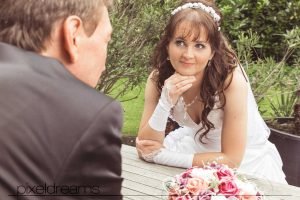 Das Brautpaar posiert an einem Tisch während der Hochzeitsfotograf ihn schräg von hinten und sie schräg von vorne Fotografiert.