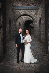 Braut und Bräutigam stehen für ihr Hochzeitsalbum auf dem Weg zum Innenhof der Burg Altena und schauen verträumt am Hochzeitsfotografen vorbei in Richtung Ausgang.