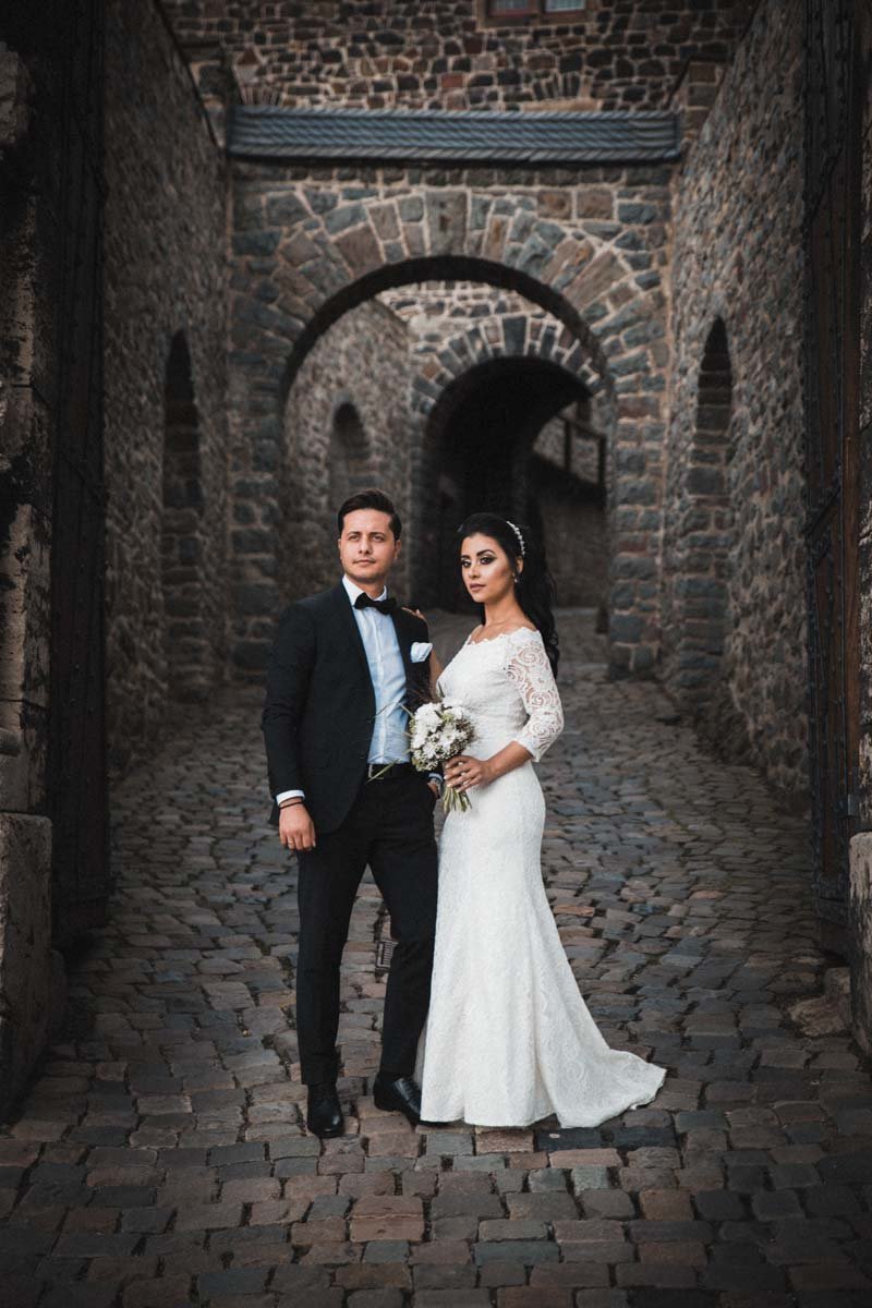 Braut und Bräutigam stehen für ihr Hochzeitsalbum auf dem Weg zum Innenhof der Burg Altena und schauen verträumt am Hochzeitsfotografen vorbei in Richtung Ausgang.