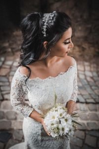 Leichte schräge Vogelperspektive der schönen Braut mit ihrem Brautstrauß. Sie schaut nach links. Im Hintergrund ein gepflasterter Weg. Schöne Brautfrisur und Brautstyling und Brautkleid.