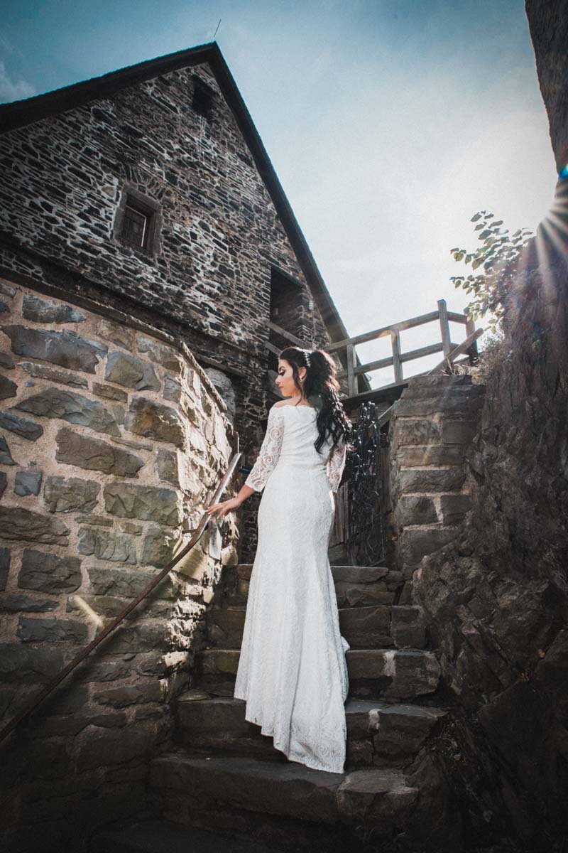 Braut steht mit dem Rücken zum Hochzeitsfotografen, auf einer Treppe in der Burg Altena, die Sonne scheint in obiger rechten Position