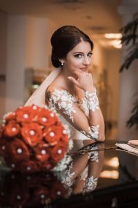 Hochzeitsfoto einer türkischen Braut Mit rotem Rosestrauß Bouquet im Vordergrund. Braut schaut lächelnd und verlegen in die Kamera. Perfekte Brautfrisur und tolles perfektes Braut Mac ab. Sie posiert über einem Klavier.